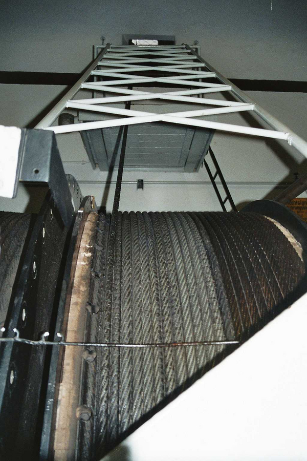 Foto: Blickrichtung von unten nach oben auf eine große Drahtseilspule unter einem weiß gestrichenen Gerüst, durch das ein Drahtseil nach oben zu einer Öffnung verläuft.
