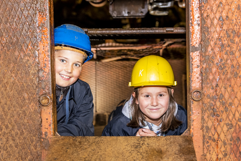 Foto: Ein Junge und ein Mädchen, bekleidet mit Schutzhelm und Overall, schauen lächelnd aus einem Waggon der Grubenbahn zum Betrachter.