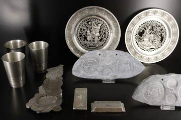 Foto zeigt den Inhalt einer Museumsvitrine mit zwei Zinntellern, zwei Zinnbarren, drei Zinnbechern, einem langen Stück geschmolzenen Zinn und zwei Gussformen für Zinnmünzen.